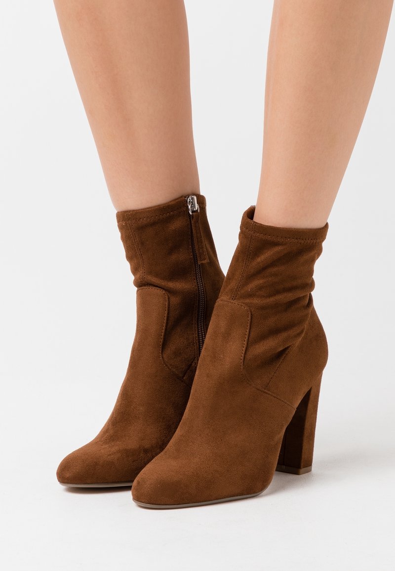 Botas de tobillo de ante marrón con tacones cuadrados y cremalleras laterales, usadas sobre piernas desnudas contra un fondo blanco.