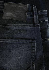 Jeans de mezclilla azul oscuro con un parche de cuero negro con texto grabado. Incluye bolsillos cosidos y detalle de botón de latón.