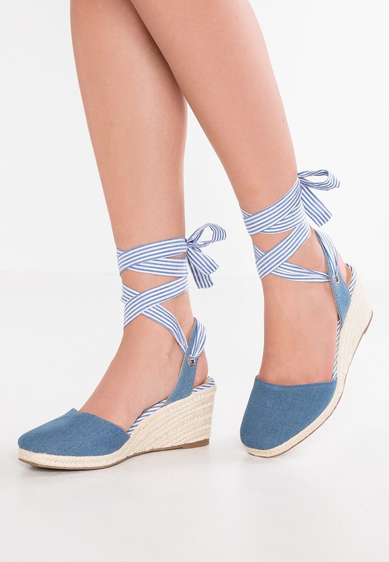 Espadrilles compensées bleues avec des liens rayés noués autour des chevilles et enroulés autour de jambes nues sur fond blanc.