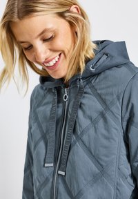 Grå vadderad jacka med huva, som har en texturerad yta, kontrasterande dragkedja och dekorativa dragband. Justerbar med en modern design.