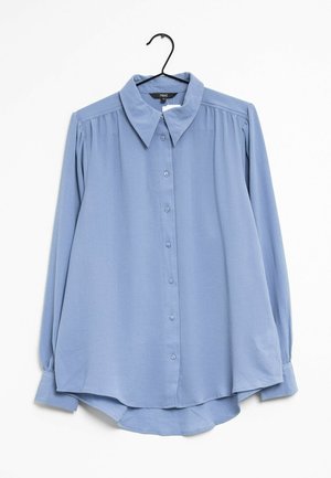 Lyseblå langærmet bluse med knapper foran, spidst krave og let rynkede skuldre, hængende på en sort bøjle.