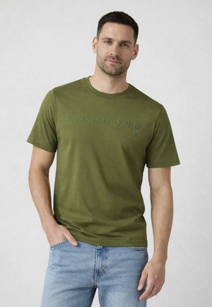 Uomo con indosso una t-shirt Richmond X verde oliva e jeans azzurri chiari, in piedi con una mano in tasca davanti a uno sfondo chiaro e semplice.