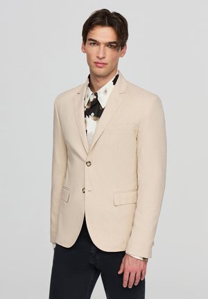 Blazer beige realizzato in tessuto testurizzato, con due tasche frontali, chiusura con un bottone singolo e bottoni a contrasto. Indossato sopra una camicia a fantasia.
