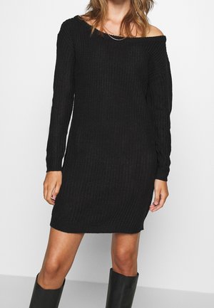 Robe tricotée noire à manches longues, design dénudé sur les épaules, texture côtelée et coupe décontractée, associée à des bottes noires hauteur genou.