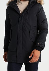 Svart parkas med en huva fodrad med päls, har en dragkedja och tryckknappslås, bröstficka och sidofickor; tillverkad av ett vattentåligt material.