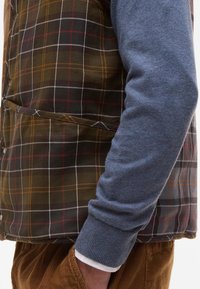 Quiltovaný kostkovaný vest v zemitých tónech hnědé a zelené, s bílými a červenými pruhy, spárovaný s pleteným svetrem v světle modré barvě.