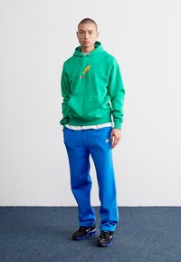 Grøn hættetrøje med et grafikdesign, parret med blå sweatpants. Sorte sportssko med et tekstureret mønster. Simpel baggrund.