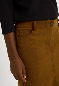 Pantalon en velours côtelé marron avec nervures verticales, une poche avant et une fermeture à bouton. Le tissu a une apparence texturée.