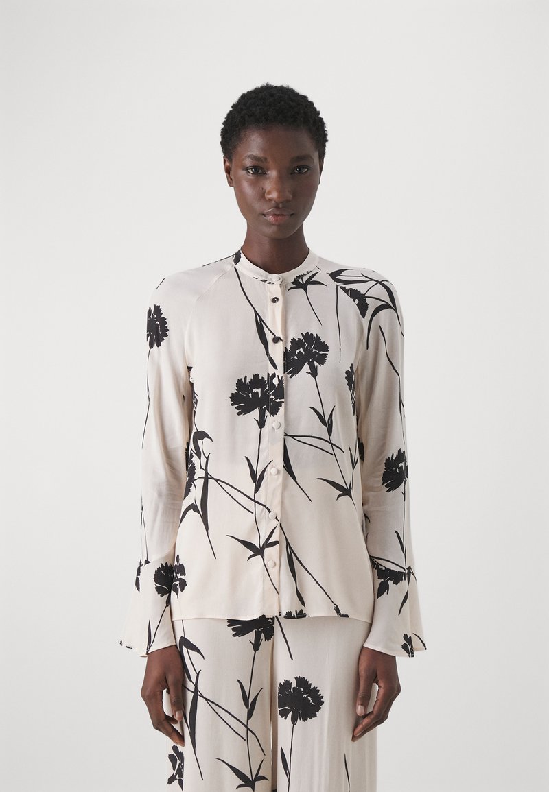 Blusa a maniche lunghe in tessuto crema chiaro con motivo floreale nero, scollatura rotonda, chiusura con bottoni e maniche a campana. Texture liscia.