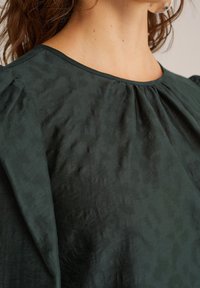 Blouse vert foncé en tissu doux et texturé, avec un col arrondi et un détail légèrement froncé aux épaules.