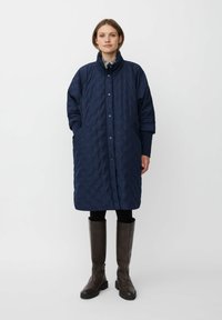 Abrigo largo acolchado de color azul marino con cuello alto, botones frontales y bolsillos laterales, combinado con botas marrones hasta la rodilla.