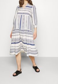 Lång, mönstrad klänning i vit tyg med blå geometriska mönster. Har V-ringning, lång ärm och en svävande nederkant. Matchas med svarta sandaler.