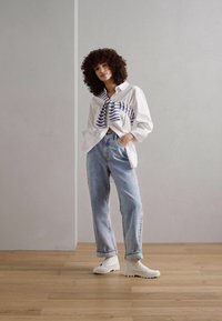 Camisa blanca de botones con rayas azules atada a la cintura, combinada con jeans holgados de color azul claro y zapatillas blancas sobre un suelo de madera.
