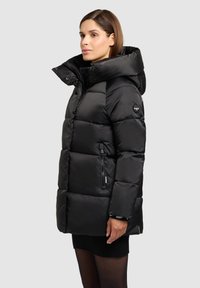 Must puffer-jope, millel on kõrge krae, luku ees ja tekstuuriga kangas. Sisaldab küljetaskut ja väikest logodetaili varrukal.