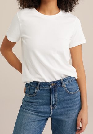 Osoba ubrana w gładką białą koszulkę z krótkim rękawem, wpuszczoną w niebieskie dżinsy z wysokim stanem, posiadające przednie kieszenie i szlufki na pasek.