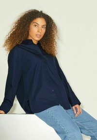 Camicia button-up blu navy a maniche lunghe con colletto, vestibilità comoda, spacco laterale e taschino sul petto, abbinata a jeans azzurri.