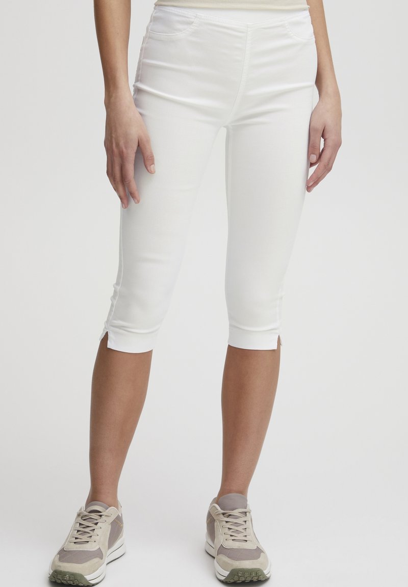 Pantalones capri blancos con un diseño ajustado, que cuentan con aberturas en los laterales en la parte inferior y bolsillos delanteros. Combinados con zapatillas deportivas de color beige claro.