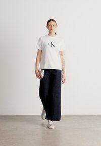 Valge puuvillane t-särk, millel on "CK" logo, kombineeritud laia säärega tumedate teksade ja hõbedaste tennistega. Modell hoiab väikest metallik-kotti.