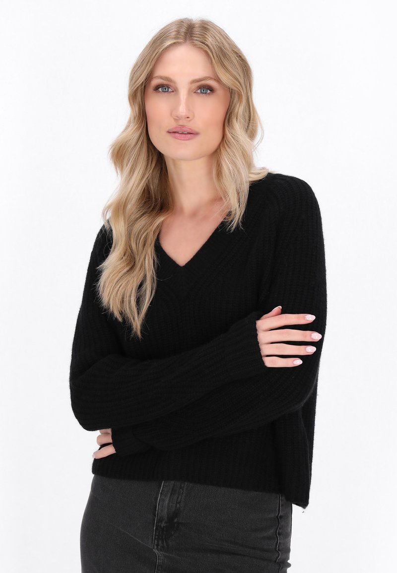 Schwarzer V-Ausschnitt-Pullover mit gerippter Textur und langen, lockeren Ärmeln. Bietet eine lässige Passform und seitliche Schlitze am Saum, kombiniert mit dunklen Jeans.