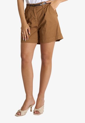 Donna che indossa pantaloncini marroni con cintura, top bianco e sandali beige con tacco e punta aperta, in piedi con una mano in tasca.
