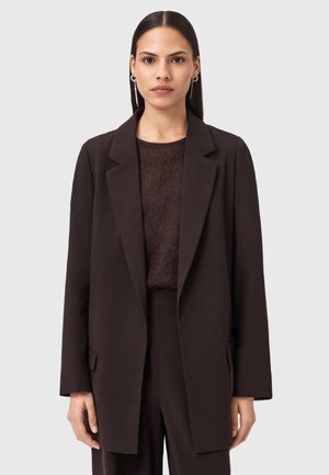 Femme avec des cheveux raides et foncés portant un long blazer marron foncé, un pantalon assorti, un pull doux marron foncé et des boucles d'oreilles en argent sur un fond neutre.