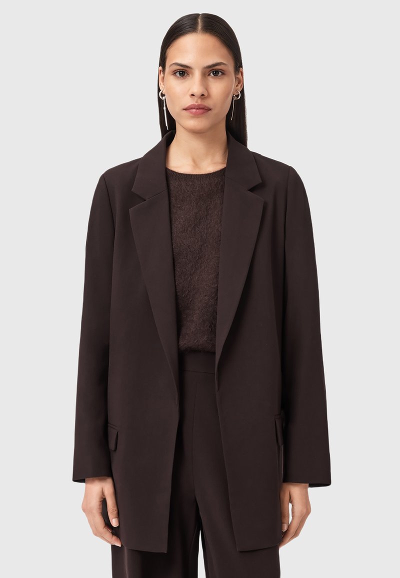 Una donna con i capelli scuri e lisci indossa un lungo blazer marrone scuro, pantaloni abbinati, un top morbido marrone scuro e orecchini d'argento, il tutto su uno sfondo neutro.