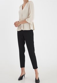 Blusa beige con scollatura a V, bottoni e design a peplum, abbinata a pantaloni neri su misura e mules neri a punta. Tessuto semplice e fluido.