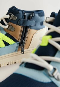 Höga sneakers med en blå, beige och ljusblå färgsättning. Funktioner inkluderar en dragkedja, texturerad tyg och kontrasterande neonaccenter.
