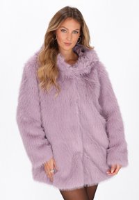 Vrouw met lang golvend haar, goudkleurige creolen en een lavendelkleurige nepbontjas met zakken, staande tegen een witte achtergrond.