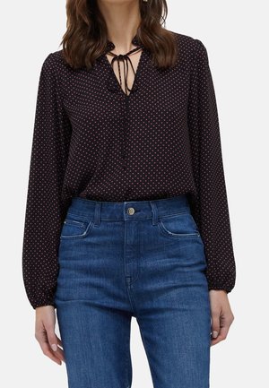 Femme portant un chemisier noir à manches longues avec de petits pois roses et un col à nouer, rentré dans un jean bleu taille haute.