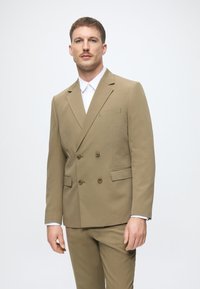 Doppelreiher beige Anzug mit zwei vorderen Taschen und braunen Knöpfen. Darunter befindet sich ein Hemd mit weißem Kragen. Glatte Textur, taillierte Passform.