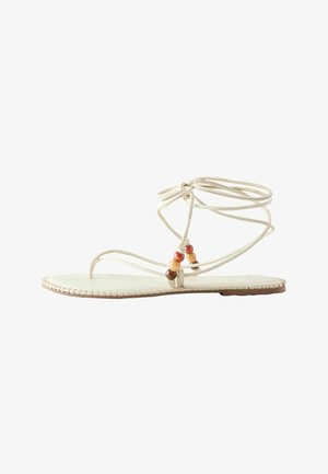 Sandalia plana de color marfil con finas tiras que envuelven el tobillo y cuentas decorativas marrones, naranjas y crema cerca del lazo del dedo.