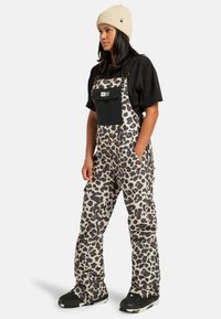 Kobieta stoi z rękami w kieszeniach, ubrana w beżowe i czarne ogrodniczki w panterkę, czarną koszulkę, beżową dzianinową czapkę i czarne trampki.