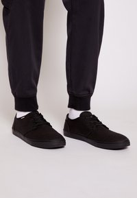 Mustat kankaiset tennarit, joissa on matala malli, teksturoitu pinta, pyöreä kärki ja kumipohja, yhdistettynä valkoisiin vartal sokkiin ja mustiin housuihin.