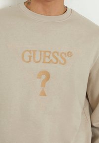 Sudadera beige con un logo en relieve de triángulo con la palabra "GUESS" y un signo de interrogación, tejido texturizado y cuello acanalado.