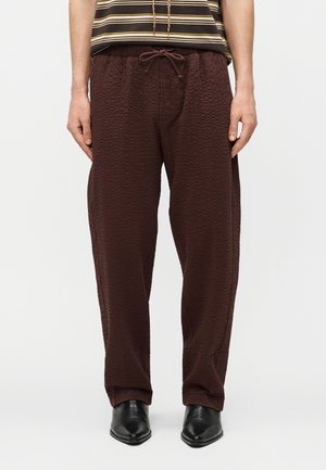 Homme portant un pantalon ample marron foncé texturé avec une taille à cordon et des chaussures pointues noires, devant un fond blanc uni.