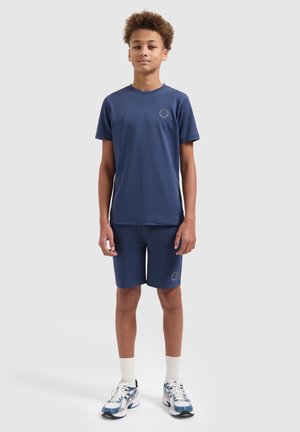 Tienerjongen staand, draagt een marineblauw shirt met korte mouwen en bijpassende korte broek, met witte sportschoenen en sokken, tegen een effen achtergrond.