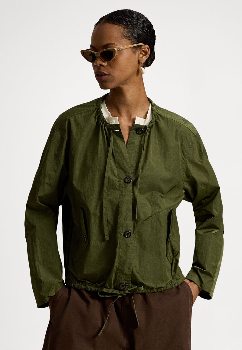 Žena v olivově zelené zapínané bundě, bílém vnitřním tričku, hnědých kalhotách, kočičí brýlemi a náušnicích ve tvaru mušlí, s rukama v kapsách.