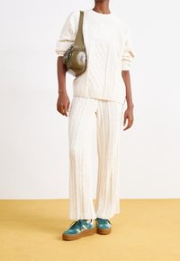 Tjock kabelstickad krämfärgad tröja och matchande vida byxor med ribbade fållar. Grön crossbody-väska och teal sneakers med bruna sulor.