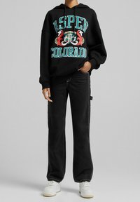 Felpa nera con grafica "ASPEN COLORADO" in turchese, rosso e bianco. Abbinata a jeans neri a gamba larga e sneakers bianche.