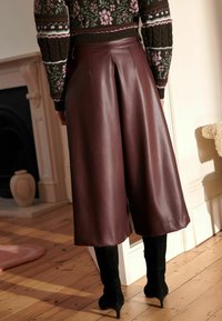 Bordeaux nepleather culottes met plooien, gecombineerd met kniehoge zwarte suède laarzen, tegen een houten vloerachtergrond.