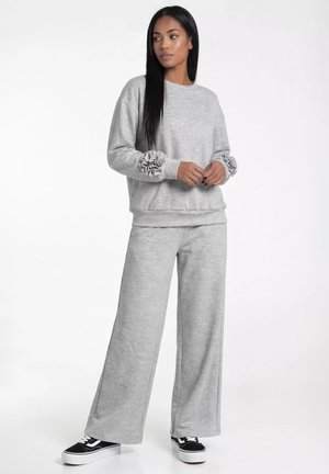 Kobieta w luźnej, jasnoszarej bluzie z falbankami na mankietach i pasujących szerokich spodniach, stojąca i lekko patrząca na bok, ubrana w czarne sneakersy.