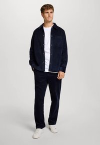 Completo in velluto a coste blu navy composto da camicia e pantaloni, abbinato a una T-shirt bianca e scarpe da ginnastica bianche. Presenta una texture a coste verticali e dettagli con bottoni.