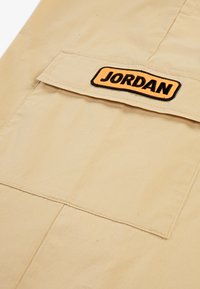 Beige stoff med en glatt tekstur, med en lapp som har "JORDAN" i svart og oransje. Inkluderer en lomme på toppen for nytte.