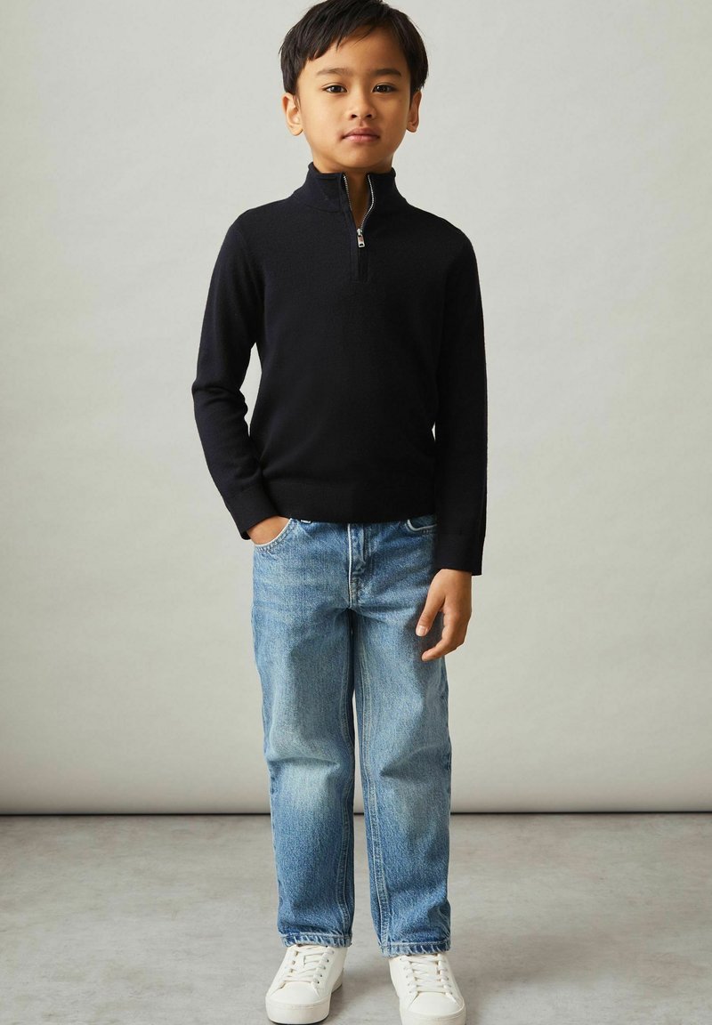 Maglione nero mezza zip, jeans in denim blu, sneakers bianche. Outfit casual con un design aderente e tessuto testurizzato.