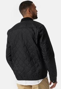 Sort quiltet jakke med bomber-stil, der har en glat tekstur og en ribbet krave. Inkluderer trykknapdetalje på mansjetten.