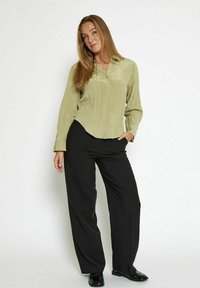 Donna con lunghi capelli biondi, che indossa una camicia button-up verde chiaro con texture, pantaloni neri a gamba larga e scarpe nere lucide, in piedi in modo rilassato.