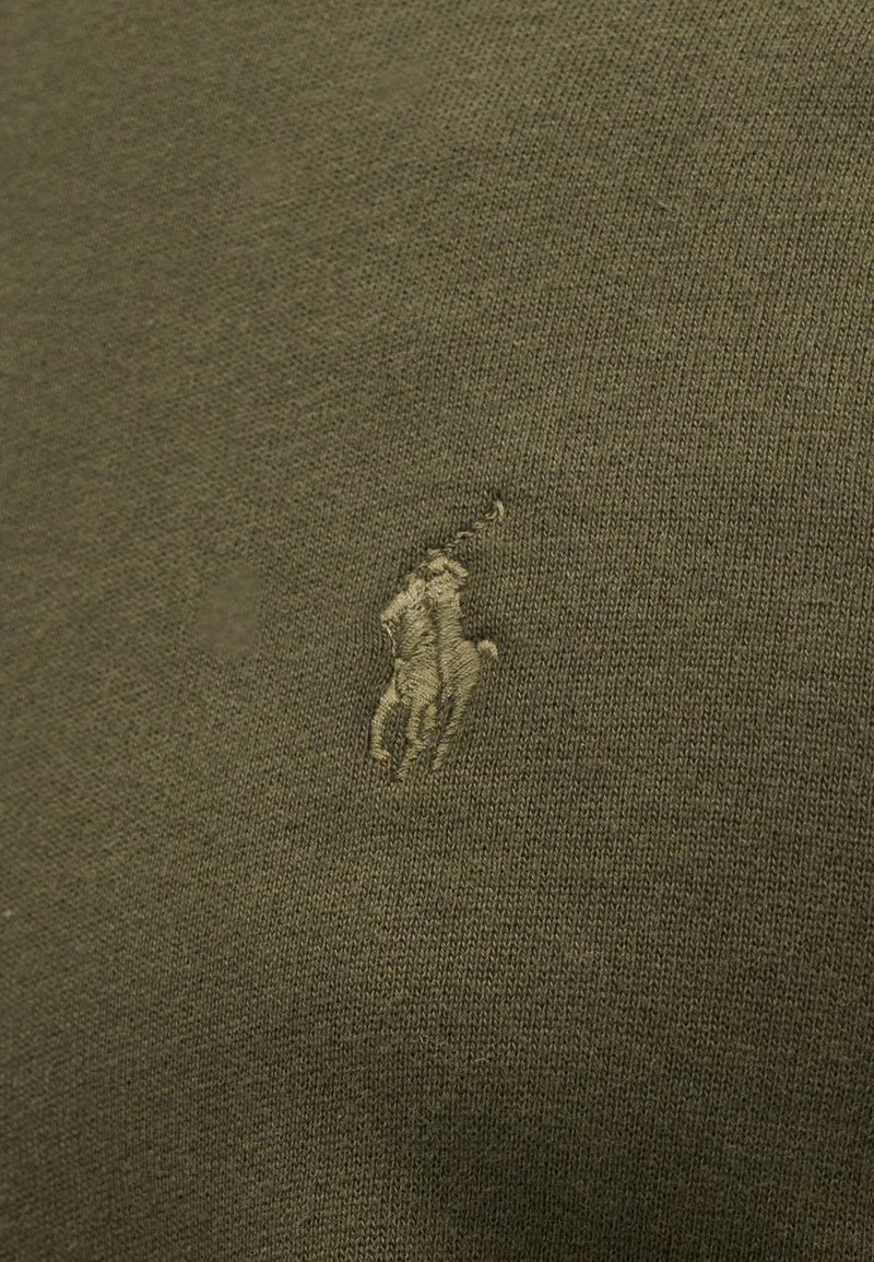 Bordado de un jugador de polo a caballo sosteniendo un mazo levantado sobre una tela de punto verde oliva.