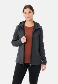 Giacca in pile grigia scura con cappuccio, tasche con zip e vestibilità aderente, indossata sopra una maglietta color ruggine, abbinata a pantaloni neri.