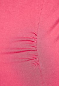 Růžová látka s hladkým povrchem, s nařasenými detaily podél jedné strany, vytvářejícími jemné rýhy a asymetrický tvar.